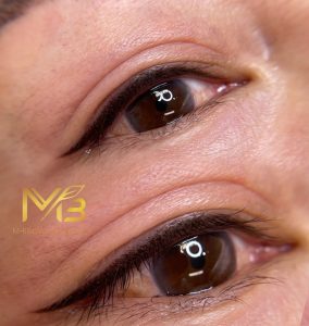 M Brows and Beauty Close-up of a person’s eyes with neatly applied black eyeliner on the upper eyelids. The skin appears smooth, and the reflection of a ring light can be seen in both eyes. The M-Brows & Beauty logo is visible in the lower left corner.