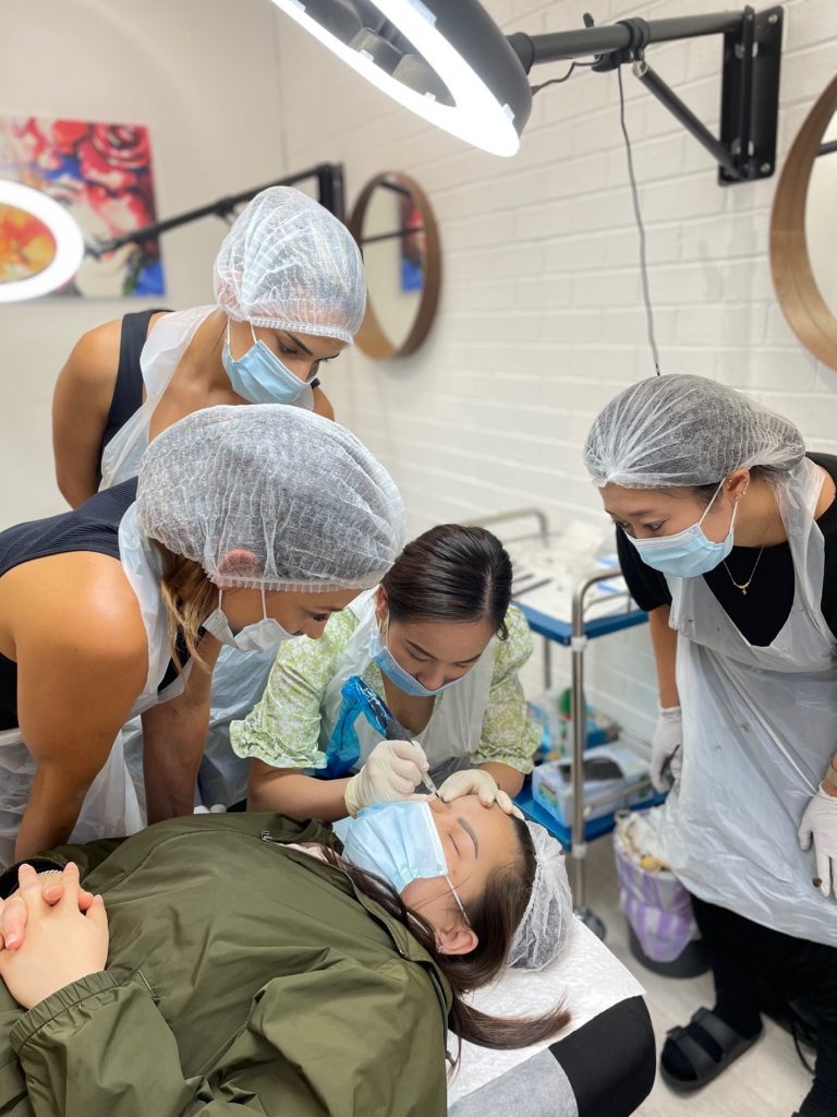 M Brows and Beauty Four women wearing hairnets, masks, and aprons closely observe a procedure as one performs a cosmetic treatment on a woman lying on a table in a clinic setting.
