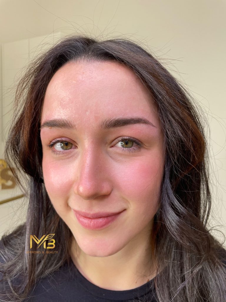 M Brows and Beauty A woman with long brown hair and green eyes smiles softly. Her cheeks and nose are slightly flushed. She wears a black shirt, and the "MB Brows & Beauty" logo hints at her expertise in cosmetic tattoo Sydney services.