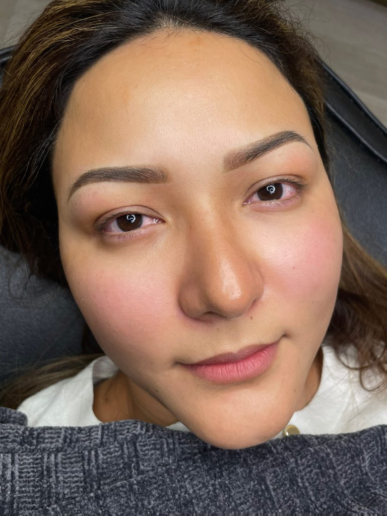 M Brows and Beauty A close-up of a woman lying down, looking up at the camera. She has well-groomed eyebrows, likely enhanced by permanent make-up, clear skin, and a neutral expression, with a textured blanket partially visible at the bottom of the image.