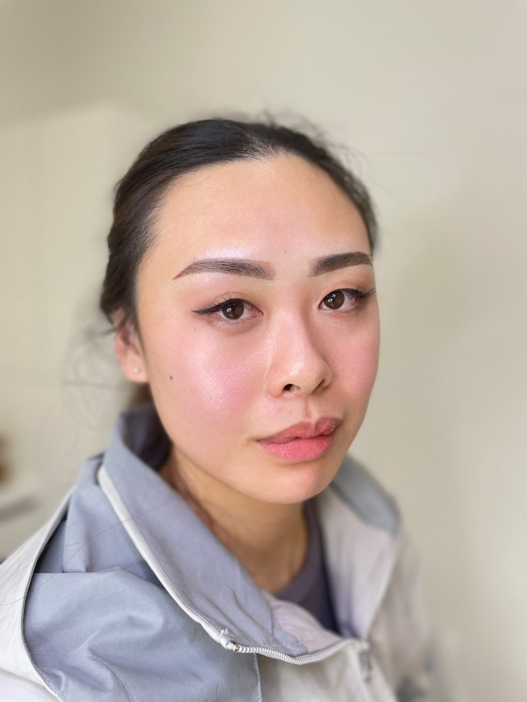 M Brows and Beauty A woman with long dark hair pulled back, showcasing expertly applied permanent make-up, wears a light grey and white jacket and faces the camera with a neutral expression against a blurred light background.