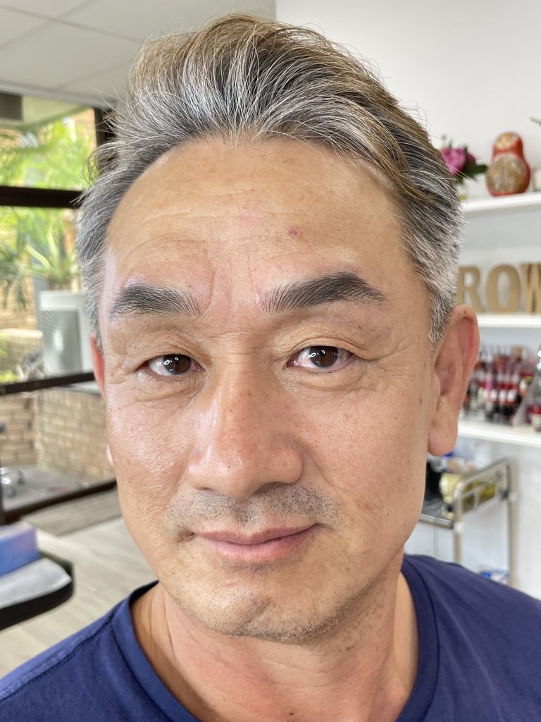 M Brows and Beauty A middle-aged man with salt-and-pepper hair and a slight smile is indoors, standing in a bright room with shelves, plants, and subtle permanent make-up enhancing his features. He is wearing a blue shirt.