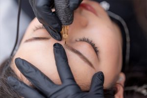 M Brows and Beauty A close-up of a person receiving a cosmetic eyebrow tattoo procedure. A gloved technician uses a tattoo machine to carefully shape and fill in the person's eyebrow whilst they keep their eyes closed.
