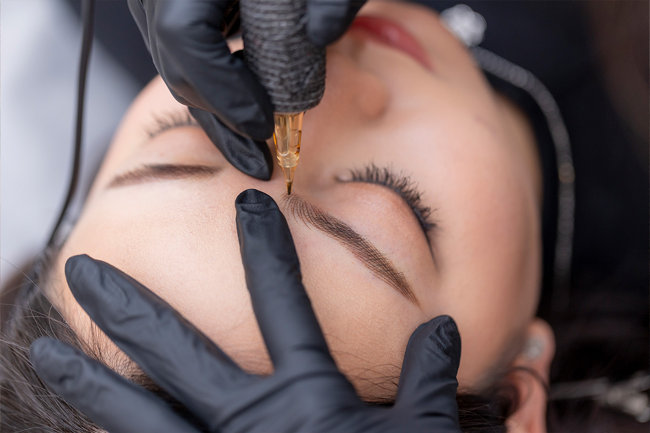 M Brows and Beauty A close-up of a person receiving a cosmetic eyebrow tattoo procedure. A gloved technician uses a tattoo machine to carefully shape and fill in the person's eyebrow whilst they keep their eyes closed.