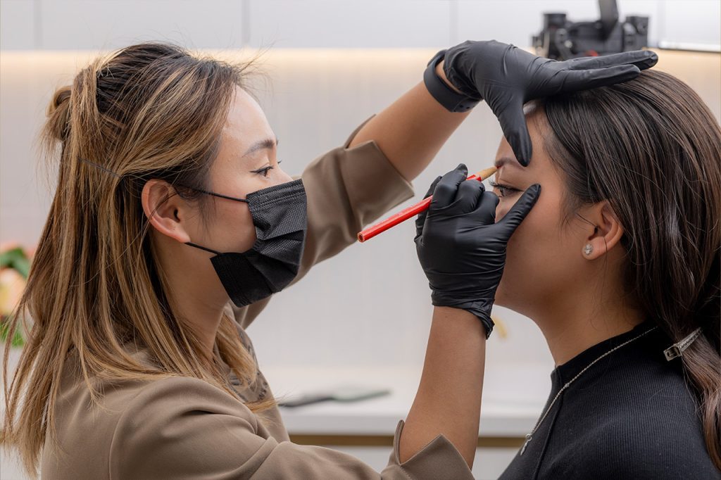 M Brows and Beauty A woman wearing a black face mask and gloves uses a red pencil to outline another woman's eyebrows in a bright, modern setting—a step in the process of applying permanent make-up. The second woman has her eyes closed and head slightly tilted back.