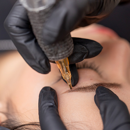 M Brows and Beauty A close-up of a person’s eyebrow being tattooed by a professional wearing black gloves, performing a cosmetic procedure with a tattoo machine.