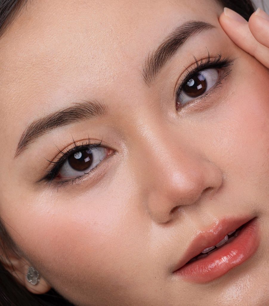 M Brows and Beauty Close-up of a Sydney woman's face with flawless skin, natural make-up, defined nano brows, long eyelashes, peachy blusher, and glossy lips. She gently touches her face, highlighting a soft and fresh look enhanced by expertly applied permanent make-up.