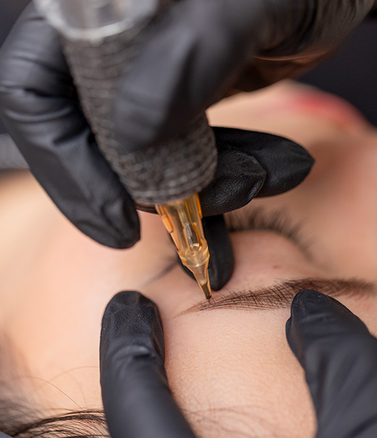 M Brows and Beauty A close-up of a person receiving an eyebrow tattoo. A gloved hand holds a tattoo machine, carefully applying permanent make-up pigment to the eyebrow area whilst another gloved hand gently stretches the skin.