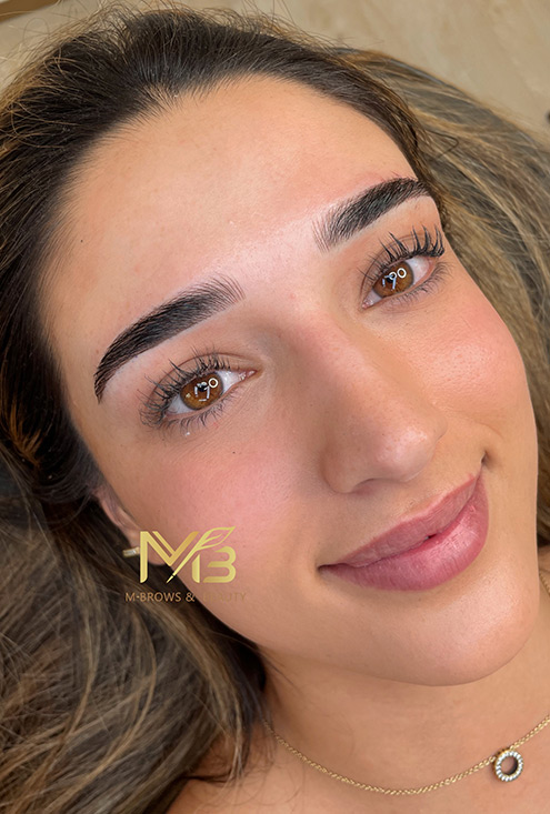 M Brows and Beauty A woman with neatly groomed dark eyebrows and long eyelashes smiles gently whilst lying down. Her skin has a natural glow, and she is wearing a delicate necklace. The MBrows & Beauty logo highlights their cosmetic tattoo training expertise.