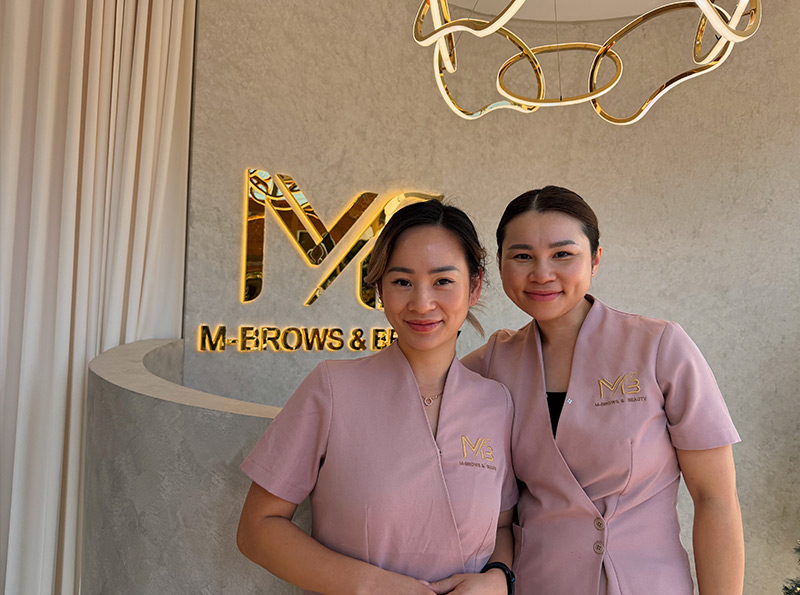 M Brows and Beauty Two women in matching pink uniforms stand smiling in front of a sign that reads "M-BROWS & BEAUTY" with a gold logo on the wall. A modern chandelier with gold rings hangs above them.
