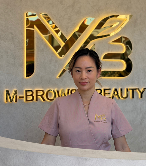 M Brows and Beauty A woman in a light purple uniform stands at a reception desk in front of a gold "M-Brows Beauty" sign on a textured wall, greeting visitors to the beauty salon.