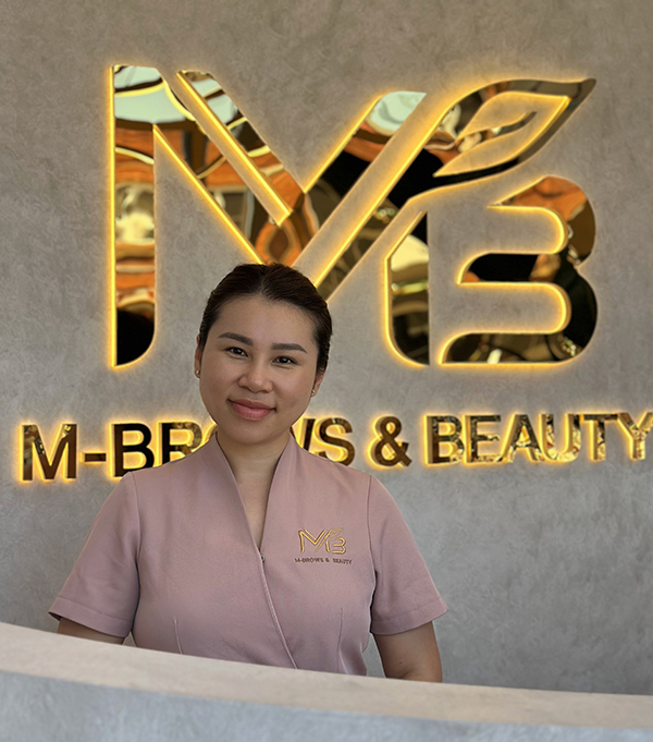 M Brows and Beauty A woman in a pink uniform stands smiling behind a white counter at M-Brows & Beauty, with a gold and black logo on the wall behind her.