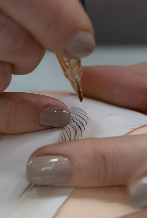 M Brows and Beauty Close-up of a hand with taupe nail varnish holding a pen or tool, drawing fine, curved lines on a surface—possibly paper or synthetic skin—while practising eyebrow microblading during cosmetic tattoo training.