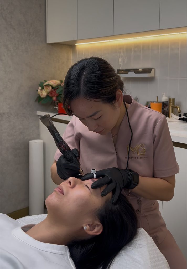 M Brows and Beauty A beauty technician in a pink uniform and black gloves tattoos a woman’s eyebrows in a modern clinic, with a flower arrangement visible in the background. The clinic also specialises in male brow tattoo treatments, including advanced nano machine hairstrokes for men seeking natural-looking results.