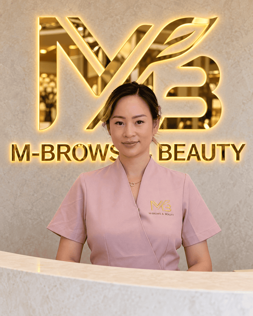 M Brows and Beauty A woman in a pink uniform stands behind a white counter, smiling slightly. Behind her, a gold “M-BROWS & BEAUTY” sign and logo are displayed on a light marble wall.
