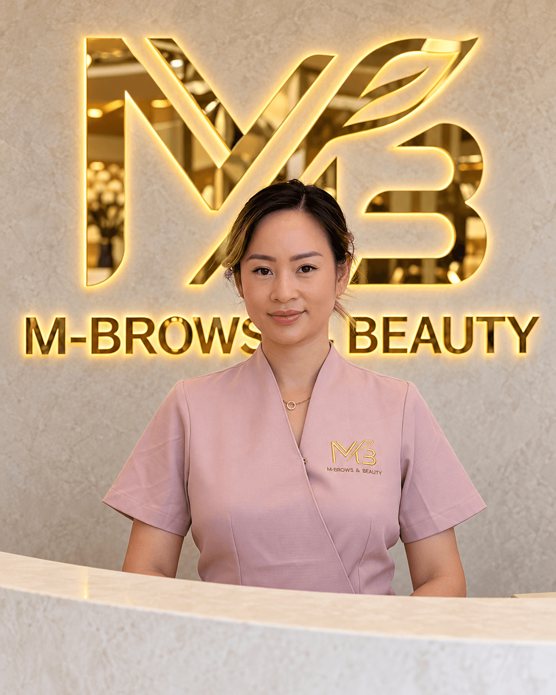 M Brows and Beauty A woman in a pink uniform stands behind a white counter, smiling slightly. Behind her, a gold “M-BROWS & BEAUTY” sign and logo are displayed on a light marble wall.