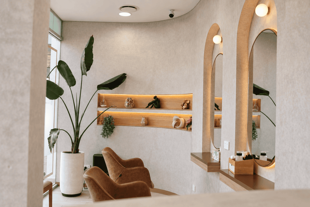 M Brows and Beauty Modern salon with two brown chairs, large potted plant, arched mirrors, and wooden shelves displaying decor items against a beige wall. Soft lighting creates a calm, inviting atmosphere.