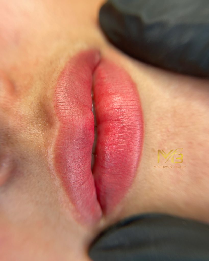 M Brows and Beauty Close-up of pink, freshly tattooed lips with gloved hands holding the face. The MBrows & Beauty logo is visible on the skin near the lips.
