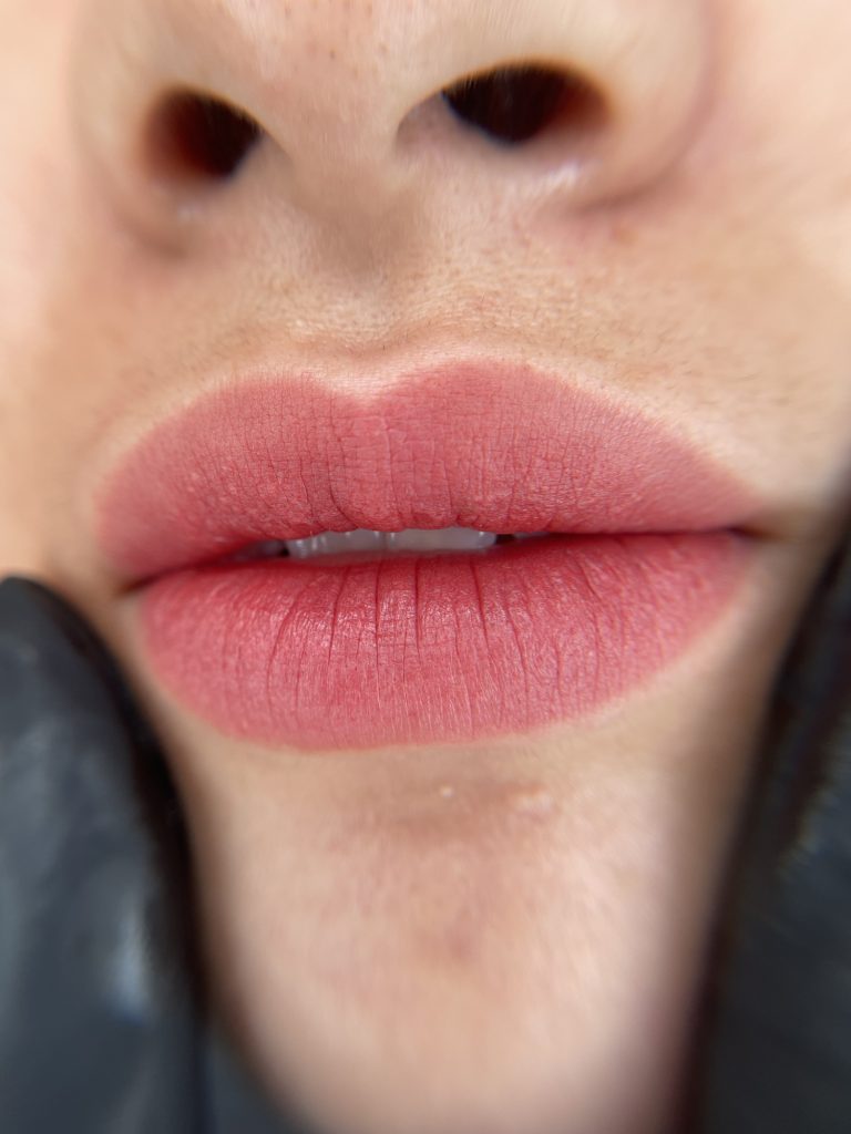 M Brows and Beauty Close-up of a person’s lips with soft pink lipstick. They have smooth skin, and a black-gloved hand gently touches their chin. The person’s nose is also partially visible at the top of the image.