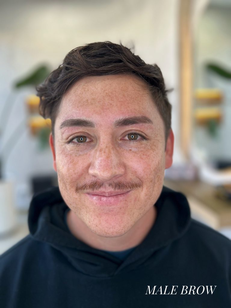 M Brows and Beauty A man with short brown hair and a trimmed moustache smiles at the camera, wearing a black hoodie. The text “MALE BROW” is written at the bottom right. The background is blurred with plants visible.