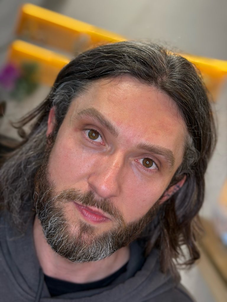 M Brows and Beauty A man with long dark hair, a full beard, and brown eyes looks at the camera. He wears a grey hoodie and is indoors with a blurred background.