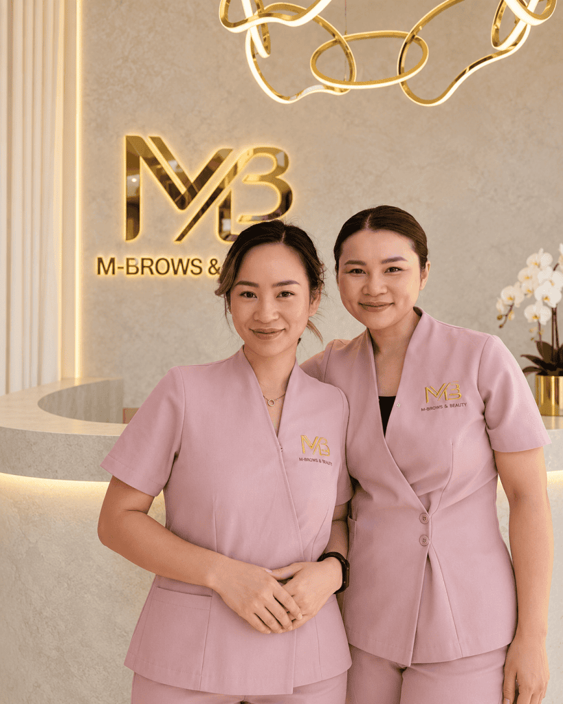 M Brows and Beauty Two women in matching pink uniforms stand and smile in front of a reception desk with a gold “M-Brows & Beauty” sign on the wall. The background includes modern décor and white orchids, highlighting the welcoming atmosphere of this renowned studio known for services such as male eyebrow tattoo Sydney, including advanced nano machine hairstrokes for men.