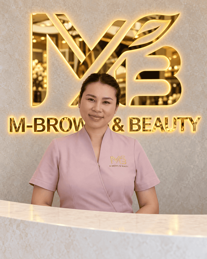M Brows and Beauty A woman in a pink uniform stands smiling behind a white counter, with a gold “M-Brows & Beauty” sign and logo on the wall in the background.