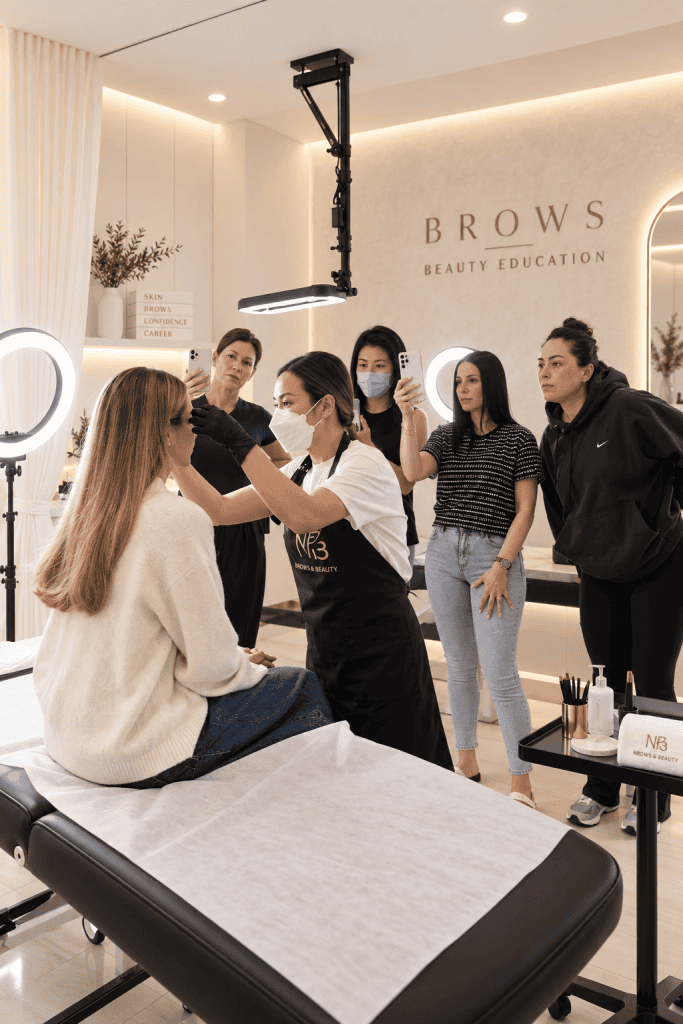 M Brows and Beauty A beauty instructor demonstrates eyebrow techniques on a seated woman whilst four students watch and record with phones in a modern, well-lit studio labelled “BROWS BEAUTY EDUCATION,” showcasing the latest trends in cosmetic tattooing Sydney.